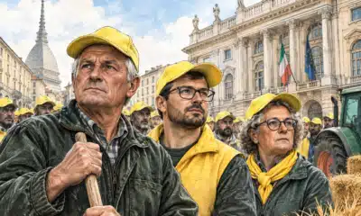 Tre agricoltori in primo piano durante una protesta, con altri manifestanti e una facciata istituzionale sullo sfondo.