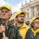 Tre agricoltori in primo piano durante una protesta, con altri manifestanti e una facciata istituzionale sullo sfondo.