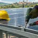 Illustrazione di pannelli fotovoltaici su una pensilina da parcheggio, con la mano guantata di un installatore in primo piano.