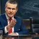 Ritratto illustrato di Pete Hegseth davanti a una sala istituzionale di pianificazione con una sedia vuota sullo sfondo.