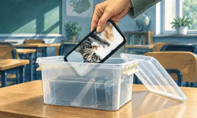 Una mano ripone uno smartphone in un contenitore su un banco, in un’aula scolastica.