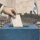 Una mano inserisce una scheda anonima in un’urna; sullo sfondo si intravede un’aula parlamentare.