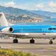 Illustrazione di un aereo regionale bianco e blu sulla pista dell’aeroporto di Genova, con il profilo costiero sullo sfondo.
