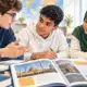 Tre studenti consultano un fascicolo didattico aperto su un banco in un’aula luminosa.