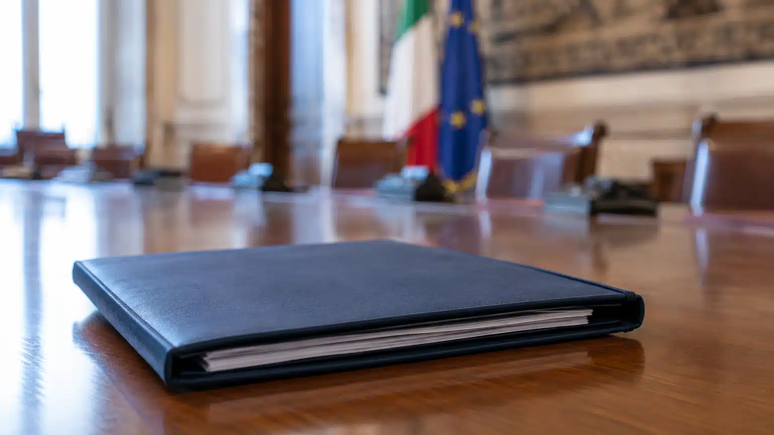 Un fascicolo di bilancio chiuso su un tavolo istituzionale, con una sala riunioni sullo sfondo