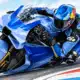 Illustrazione di un pilota con casco e tuta blu su una moto blu inclinata in curva.