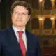 Ritratto illustrato di un uomo in abito scuro davanti a un interno teatrale appena accennato.