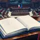 Un fascicolo legislativo aperto su un banco, con l’aula parlamentare sullo sfondo illustrato.