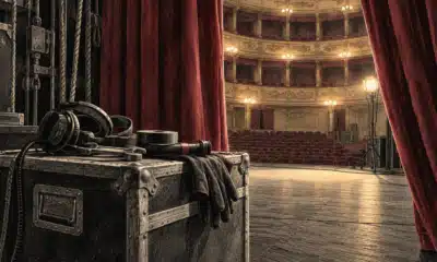 Cuffia e attrezzi da macchinista su un baule in quinta, con il palcoscenico illuminato oltre il sipario.