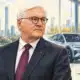 Ritratto illustrato di Frank-Walter Steinmeier davanti a un ambiente simbolico di ricerca automobilistica, con una sagoma di veicolo elettrico sullo sfondo.