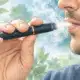 Una mano porta alla bocca un dispositivo per tabacco riscaldato, in un’illustrazione dai toni verde salvia e azzurro.