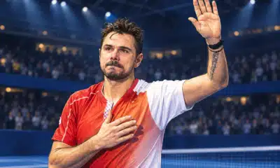 Un tennista ispirato alla foto di riferimento saluta il pubblico in un’arena indoor, con una mano alzata e l’altra sul petto.