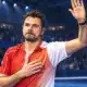 Un tennista ispirato alla foto di riferimento saluta il pubblico in un’arena indoor, con una mano alzata e l’altra sul petto.